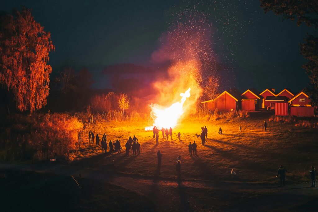 A large group enjoys a bright bonfire at night in a rural village setting, creating a warm and communal atmosphere.